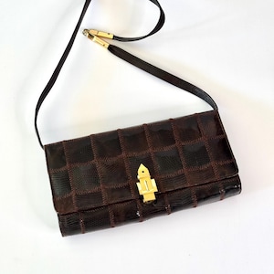 Può includere: Una borsa marrone vintage con un motivo a scacchi e una lunga tracolla. La borsa presenta una chiusura e dettagli color oro. La tracolla è sottile e marrone scuro. La borsa è rettangolare.
