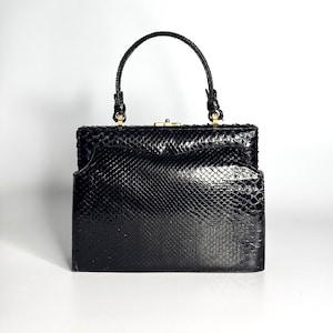Pode incluir: Uma bolsa de mão preta estruturada com um padrão texturizado de pele de cobra. A bolsa possui uma alça superior, um fecho de metal dourado no topo e uma silhueta retangular rígida com um detalhe de aba ligeiramente curvada na frente.