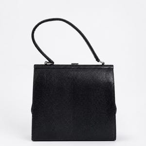 Könnte beinhalten: Eine schwarze Handtasche mit strukturierter, quadratischer Silhouette und einer Textur in Eidechsenoptik. Die Tasche verfügt über einen einzelnen, gebogenen Tragegriff mit silberfarbenen Metallbeschlägen und einen einfachen Metallverschluss.