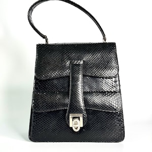 Könnte beinhalten: Eine schwarze Handtasche mit texturiertem Schlangenledermuster, ausgestattet mit einem einzelnen Tragegriff, einer Umschlagklappe und einem zentralen vertikalen Riemen, der mit einem silberfarbenen Metalldrehverschluss gesichert ist.