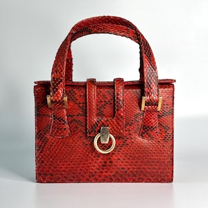 Könnte beinhalten: Eine strukturierte Handtasche mit einem leuchtend roten und schwarzen Schlangenleder-Muster. Die Tasche verfügt über einen einzelnen Tragegriff, eine rechteckige Form und einen zentralen vertikalen Riemenverschluss mit einem goldenen Metallring.