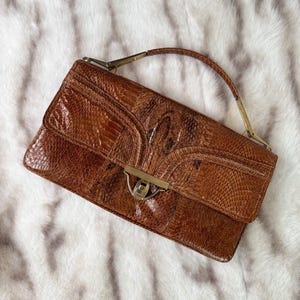 Puede incluir: Bolso de mano vintage de piel de serpiente marrón con asa superior y cierre dorado. El bolso rectangular presenta un patrón texturizado y un diseño estructurado. El bolso está sobre un fondo blanco y esponjoso.