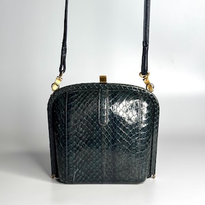 Könnte beinhalten: Eine dunkelgrüne, strukturierte Handtasche mit schlangenlederartiger Textur, ausgestattet mit einem goldfarbenen Metallverschluss oben in der Mitte und einem dünnen, passenden Schulterriemen mit goldfarbenen Beschlägen.