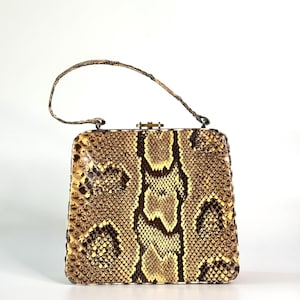 Puede incluir: Un bolso vintage con un estampado de piel de serpiente en tonos marrones y beige. El bolso rectangular tiene un asa corta y un cierre dorado. El estampado de piel de serpiente se muestra prominentemente en la parte delantera y los lados del bolso.