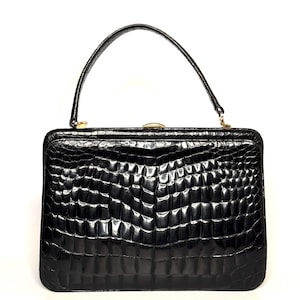 Könnte beinhalten: Eine strukturierte schwarze Handtasche mit glänzender, texturierter Oberfläche und einem ausgeprägten schuppenartigen Muster. Die Tasche verfügt über einen einzelnen, gebogenen Tragegriff und einen goldfarbenen Metallverschluss in der Mitte.