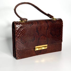 Può includere: Una borsa a mano marrone in pelle di serpente con manico superiore e chiusura dorata. La borsa rettangolare ha una superficie strutturata e una chiusura a patta. Il manico è curvo e fissato alla parte superiore della borsa con finiture dorate.