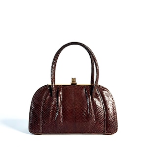 May include: A dark brown handbag with a textured snakeskin pattern, featuring a structured semi-circular shape, two matching top handles, and a gold-toned metal frame closure at the top center. The bag has a polished finish and pleated details.