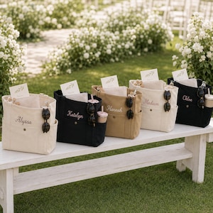 May include: Five canvas tote bags in cream, black, and tan colors, each featuring a personalized embroidered name on the front pocket. The bags include side pockets holding sunglasses and tumblers, with small cards tucked inside the main compartments.