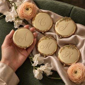 May include: Five round gold-toned metal compact mirrors, each featuring a single engraved cursive letter. The letters displayed are A, C, J, M, and R. The mirrors are arranged on a soft, draped champagne-coloured fabric with peach ranunculus flowers.