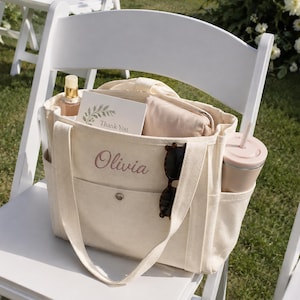 May include: A cream-colored canvas tote bag featuring the name Olivia embroidered in pink script on the front pocket. The bag includes side pockets, a snap closure, and contains a Thank You card, a beige pouch, sunglasses, and a tumbler.