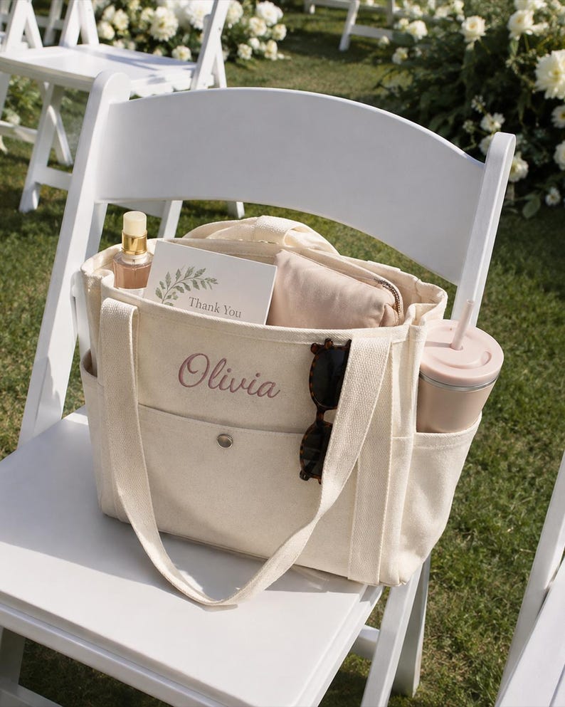 May include: A cream-colored canvas tote bag featuring the name Olivia embroidered in pink script on the front pocket. The bag includes side pockets, a snap closure, and contains a Thank You card, a beige pouch, sunglasses, and a tumbler.