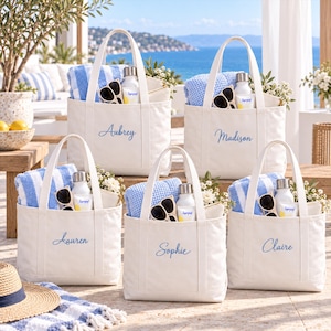 Pode incluir: Cinco sacos de lona em tom branco sujo com duas alças, cada um bordado com um nome em letra azul: Aubrey, Madison, Lauren, Sophie e Claire. Cada saco contém uma toalha às riscas azuis e brancas, uma garrafa de água branca e óculos de sol.