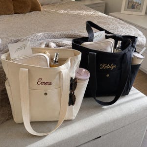 May include: Two canvas tote bags, one cream and one black, each featuring a front pocket with a snap button and embroidered names Emma and Katelyn. The bags contain small zippered pouches, spray bottles, and side pockets holding tumblers.