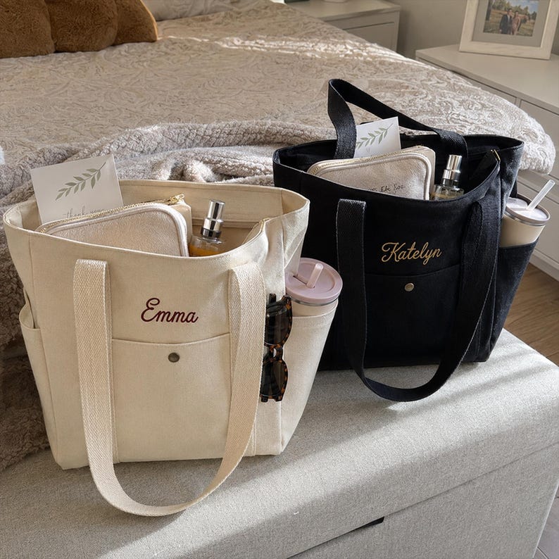 May include: Two canvas tote bags, one cream and one black, each featuring a front pocket with a snap button and embroidered names Emma and Katelyn. The bags contain small zippered pouches, spray bottles, and side pockets holding tumblers.