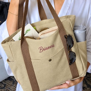 May include: A beige canvas tote bag with brown webbing handles and a front pocket featuring the embroidered name Brianna in dark red script. The bag contains a small pouch, a card, and a grey tumbler, with tortoiseshell sunglasses attached.