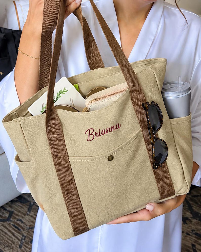 May include: A beige canvas tote bag with brown webbing handles and a front pocket featuring the embroidered name Brianna in dark red script. The bag contains a small pouch, a card, and a grey tumbler, with tortoiseshell sunglasses attached.