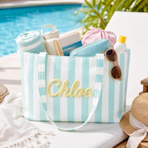 Puede incluir: Un bolso de lona a rayas azul claro y blanco con el nombre Chloe bordado en letra cursiva amarilla en la parte delantera. El bolso contiene una toalla enrollada, un libro, una bolsa rosa, un estuche azul, una botella blanca y gafas de sol.