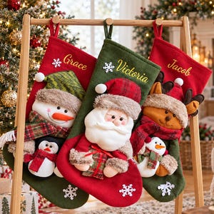 May include: Three Christmas stockings hanging on a wooden stand. The left is red with a snowman and penguin, the center is green with Santa Claus, and the right is red with a reindeer and snowman. Each features embroidered names Grace, Victoria, and Jacob.
