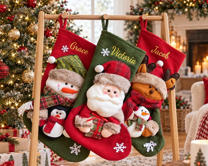 May include: Three Christmas stockings hanging on a wooden stand. The left is red with a snowman and penguin, the center is green with Santa Claus, and the right is red with a reindeer and snowman. Each features embroidered names Grace, Victoria, and Jacob.