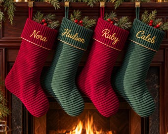 May include: Four ribbed Christmas stockings hanging in alternating red and dark green colors. Each stocking features a gold-trimmed cuff embroidered with the names Nora, Hudson, Ruby, and Caleb in gold script. Pine sprigs and red berries fill the tops.