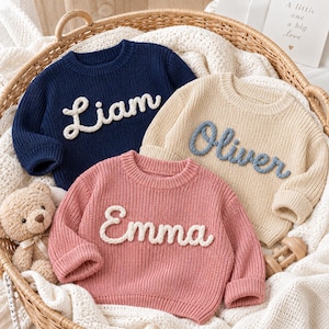 May include: Three long-sleeve knit sweaters in navy blue, beige, and dusty pink. Each sweater features a crew neckline and a textured, raised script name appliqued on the front in white or blue yarn. The names read Liam, Oliver, and Emma.