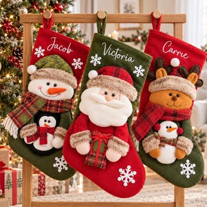 May include: Three Christmas stockings hanging on a wooden stand. The left is red and green with a snowman and penguin. The center is green and red with a Santa Claus. The right is red and green with a reindeer and snowman. Each has a name embroidered.