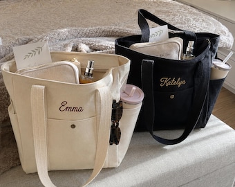 May include: Two canvas tote bags, one cream and one black, each featuring a front pocket with a snap button and an embroidered name. The cream bag is embroidered with the name Emma, and the black bag is embroidered with the name Katelyn.