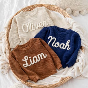 May include: Three long-sleeved knit jumpers in cream, navy blue, and brown. Each jumper features a crew neckline and ribbed cuffs. The cream jumper has the name Oliver, the navy blue jumper has the name Noah, and the brown jumper has the name Liam.