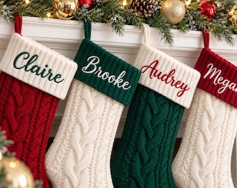 May include: Four cable-knit Christmas stockings hanging in a row. The first is red with a white cuff embroidered with the name Claire. The second is white with a green cuff embroidered with Brooke. The third is green with a white cuff embroidered with Audrey. The fourth is white with a red cuff embroidered with Megan.