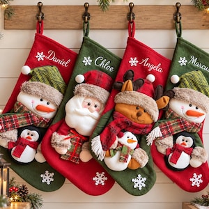 May include: Four Christmas stockings in red and green felt, each featuring a plush character: a snowman with a penguin, Santa Claus, a reindeer with a snowman, and another snowman with a penguin. Each stocking is embroidered with a name.