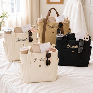 May include: Four canvas tote bags in cream, tan, and black, each personalized with embroidered names: Amanda, Sophie, Rachel, and Danielle. Each bag features dual handles, side pockets, a front snap closure, and contains a tumbler, pouch, and sunglasses.