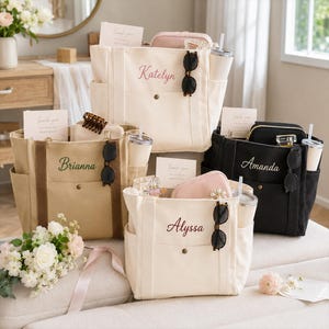 May include: Four canvas tote bags in cream, tan, and black colors, each personalized with embroidered names including Katelyn, Brianna, Amanda, and Alyssa. Each bag features side pockets, a front snap closure, sunglasses, and a tumbler.