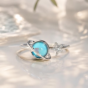 May include: A silver-toned open ring featuring a central blue spherical charm encircled by a thin ring to resemble a planet, paired with a small five-pointed star charm encrusted with clear stones on the opposite end of the adjustable band.