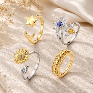May include: Four adjustable rings with nature-inspired designs. One gold ring features a star and crescent moon. Another has a sunflower, and a third has a beaded band. The fourth ring has a flower with a blue center and yellow gem.