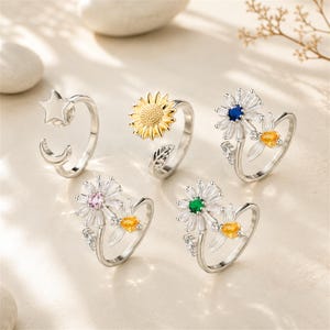 May include: A collection of five silver-toned open rings. One features a star and crescent moon. Another is a sunflower with a leaf. Three rings feature daisy-shaped designs with white petals and central stones in blue, pink, and green, each paired with a small bee.
