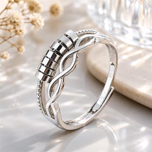 May include: A silver-toned ring featuring a polished finish with a series of five small, cylindrical beads that rotate along the top section. The band incorporates an intricate, twisted crossover design and a beaded edge detail along one side.