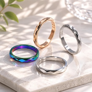 May include: Four rings with geometric diamond-cut designs. One ring is iridescent with blue, green, and purple hues. The other rings are gold, silver, and stainless steel. The rings are displayed on a white surface.