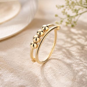 May include: A gold-colored ring with a double band design. The top band features four small, spherical beads. The ring is made of a shiny, reflective metal and is set against a soft, neutral background. The ring is a piece of jewelry.