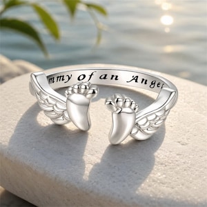 May include: A silver ring with the inscription "Mommy of an Angel". The ring features two baby feet and angel wings on either side. The ring is displayed on a light-colored, textured surface.