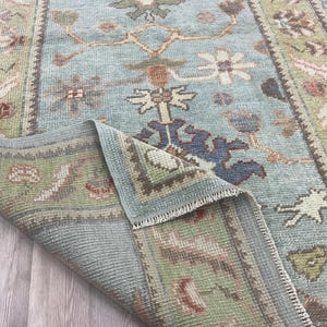 May include: A light blue area rug featuring a repeating floral and geometric pattern in shades of sage green, rust orange, and dark blue. The rug has a folded corner revealing the underside, showcasing a woven texture and a fringed edge detail.
