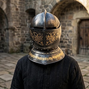 May include: A dark metal knight helmet featuring a pointed crown, a horizontal visor slit, and intricate gold-colored filigree engravings on the faceplate and neck guard. The helmet is worn over a black quilted garment with a textured fabric surface.