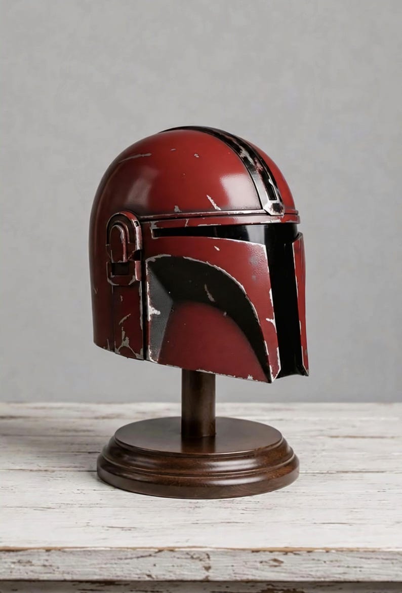 May include: A dark red helmet with a T-shaped visor and a weathered, distressed finish featuring silver-colored scratches and scuffs. The helmet is mounted on a dark brown, circular wooden pedestal stand with a central vertical support column.