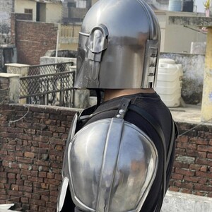 May include: A metallic silver helmet with a T-shaped visor and a matching shoulder pauldron piece. The helmet features a smooth, rounded dome and vertical vents on the side. The shoulder armor is curved and polished, secured with black straps.