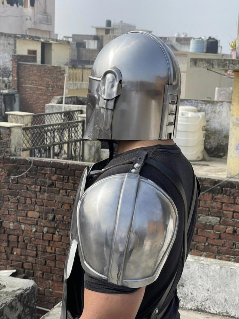 May include: A metallic silver helmet with a T-shaped visor and a matching shoulder pauldron piece. The helmet features a smooth, rounded dome and vertical vents on the side. The shoulder armor is curved and polished, secured with black straps.