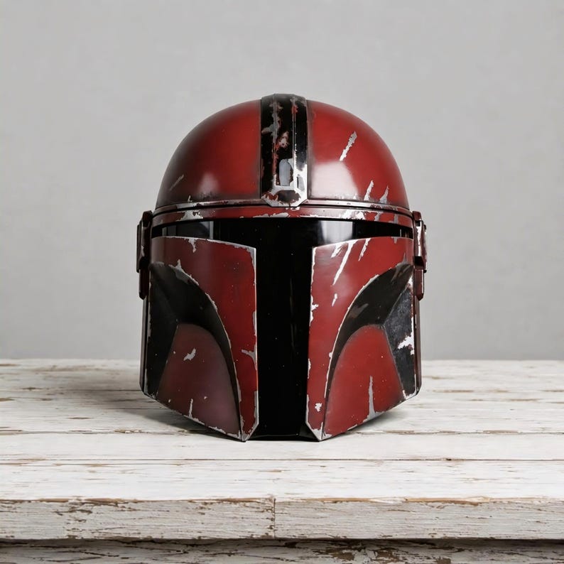 May include: A red Mandalorian-style helmet featuring a black T-shaped visor and a weathered, distressed finish with visible silver-colored scratches and paint chips across the surface, showcasing a rugged, battle-worn appearance throughout.