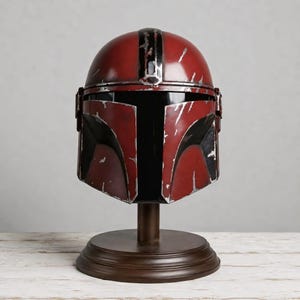 May include: A red and black helmet with a weathered, distressed finish featuring silver-colored scratches and scuffs across the surface. The helmet has a T-shaped visor opening and is mounted on a circular brown pedestal stand for display purposes.