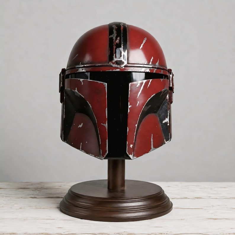 May include: A red and black helmet with a weathered, distressed finish featuring silver-colored scratches and scuffs across the surface. The helmet has a T-shaped visor opening and is mounted on a circular brown pedestal stand for display purposes.