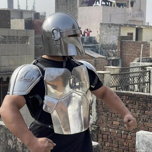 May include: A metallic silver helmet with a T-shaped visor and matching chest and shoulder armor plates. The armor features a polished finish and is secured with black leather straps over a plain black t-shirt and light-colored pants.