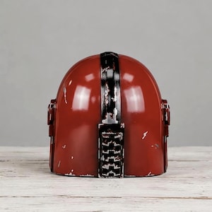 May include: A glossy red helmet featuring a central black vertical stripe with a distressed, weathered finish. The helmet has a rounded dome shape with small rectangular side attachments and visible silver-toned scuffs along the edges.