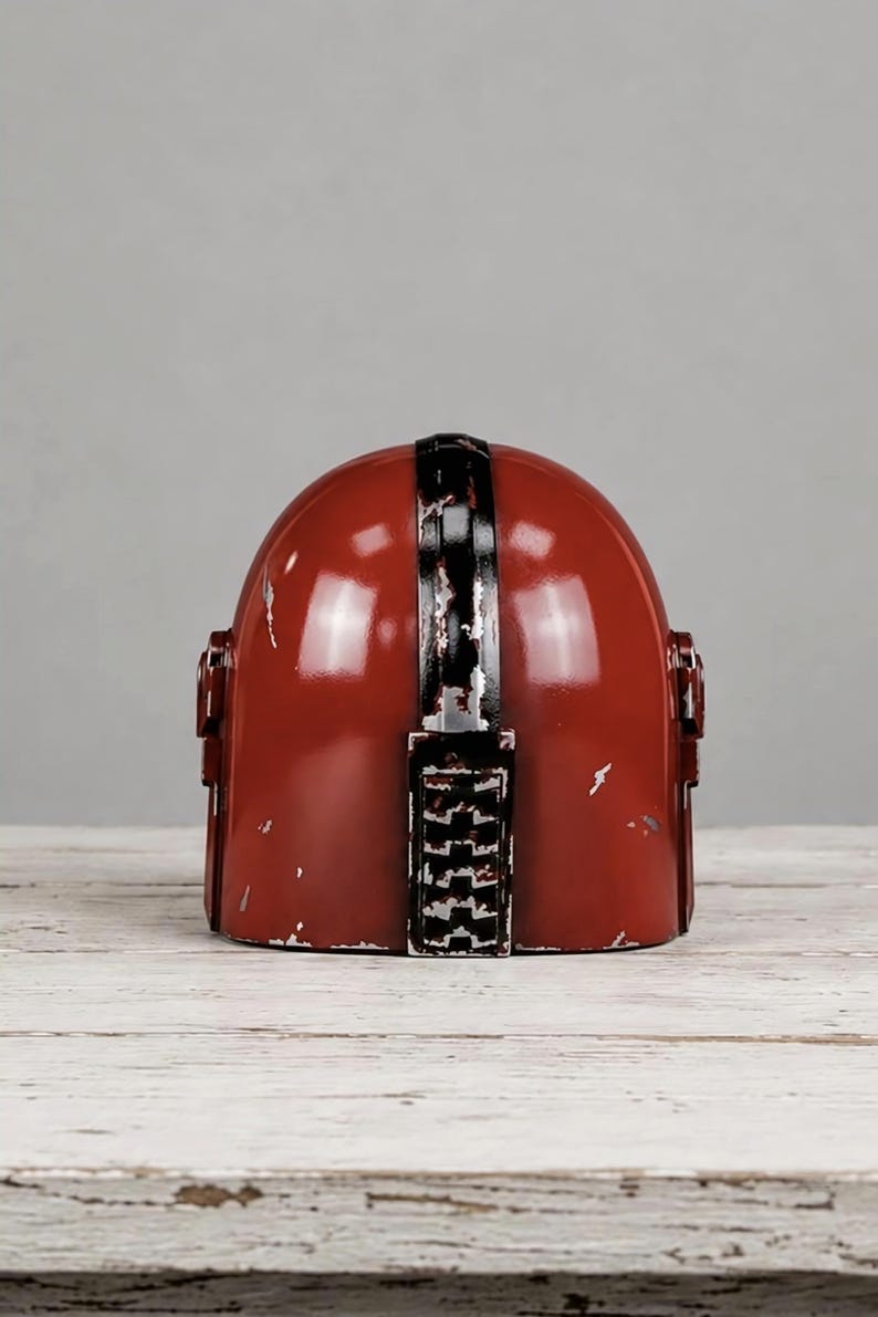May include: A glossy red helmet featuring a central black vertical stripe with a distressed, weathered finish. The helmet has a rounded dome shape with small rectangular side attachments and visible silver-toned scuffs along the edges.